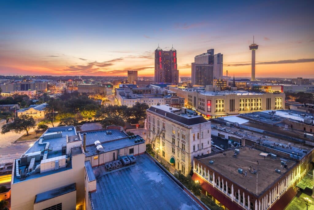 An elevated panoramic view of the San Antonio, Texas skyline at dusk, featuring the Tower of the Americas and diverse urban architecture under a vibrant sunset.