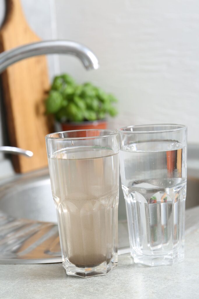 Two glasses of water sit on a kitchen counter near a sink, with one containing murky, brown water and the other containing clear water.