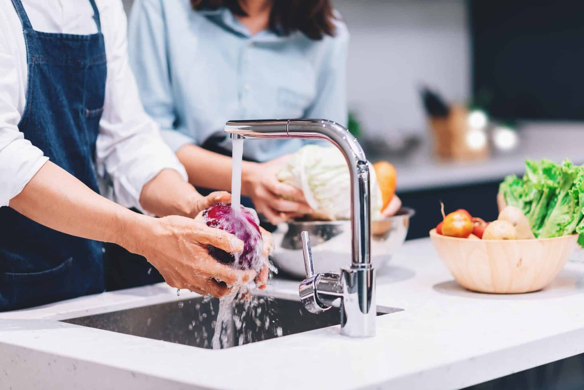 A person in an apron washes a head of red cabbage under a running chrome faucet in a modern kitchen while another person prepares a green cabbage in the background.