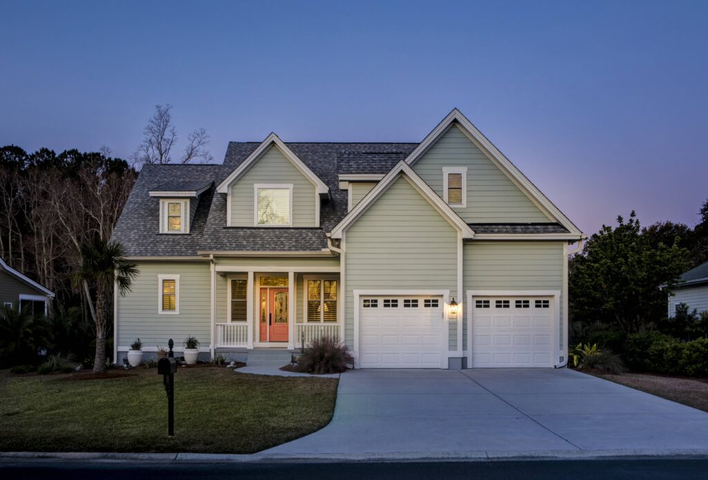 A two-story, light green suburban home with white trim and a two-car garage is illuminated against a deep blue dusk sky.