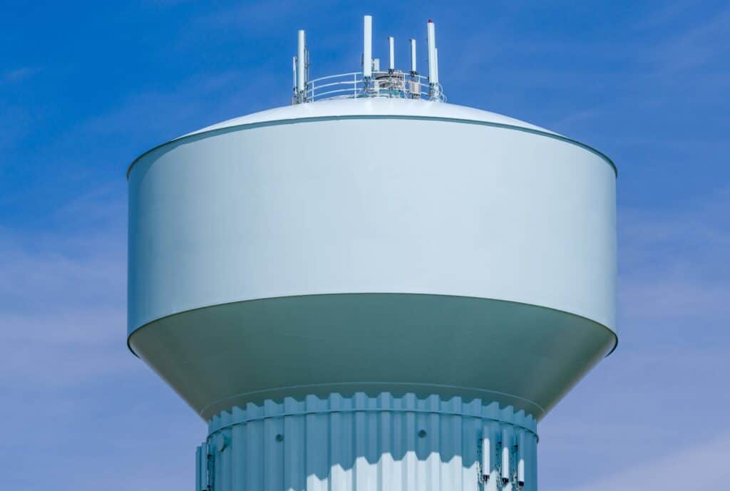 A large, pale blue modern water tower with multiple communication antennas on top stands against a clear, bright blue sky.