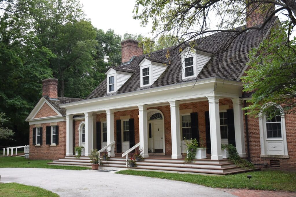 This historic, red brick colonial-style mansion features a grand white-columned portico, symmetrical windows with black shutters, and three pedimented dormer windows atop a shingle roof.