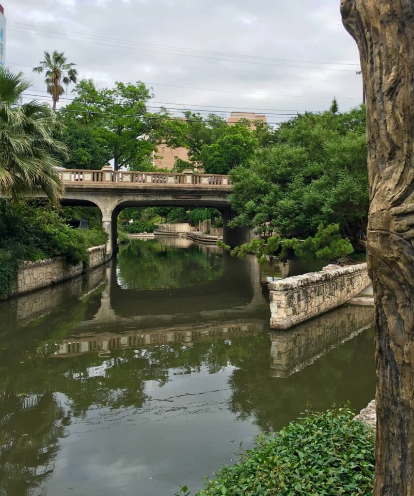 A peaceful view of a calm river flowing under a concrete bridge, surrounded by lush green trees and stone-lined banks under an overcast sky.