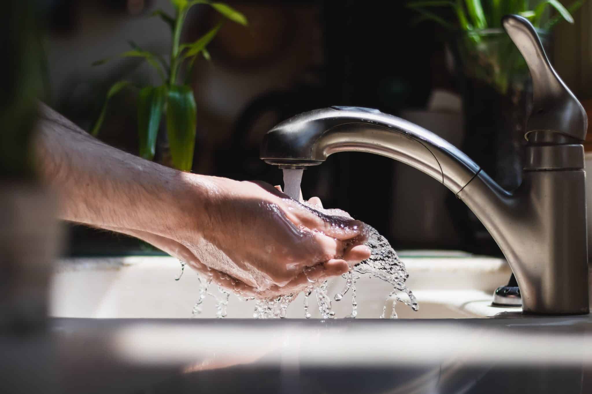 A close-up shot shows a person washing their hands under a stream of water from a chrome kitchen faucet.