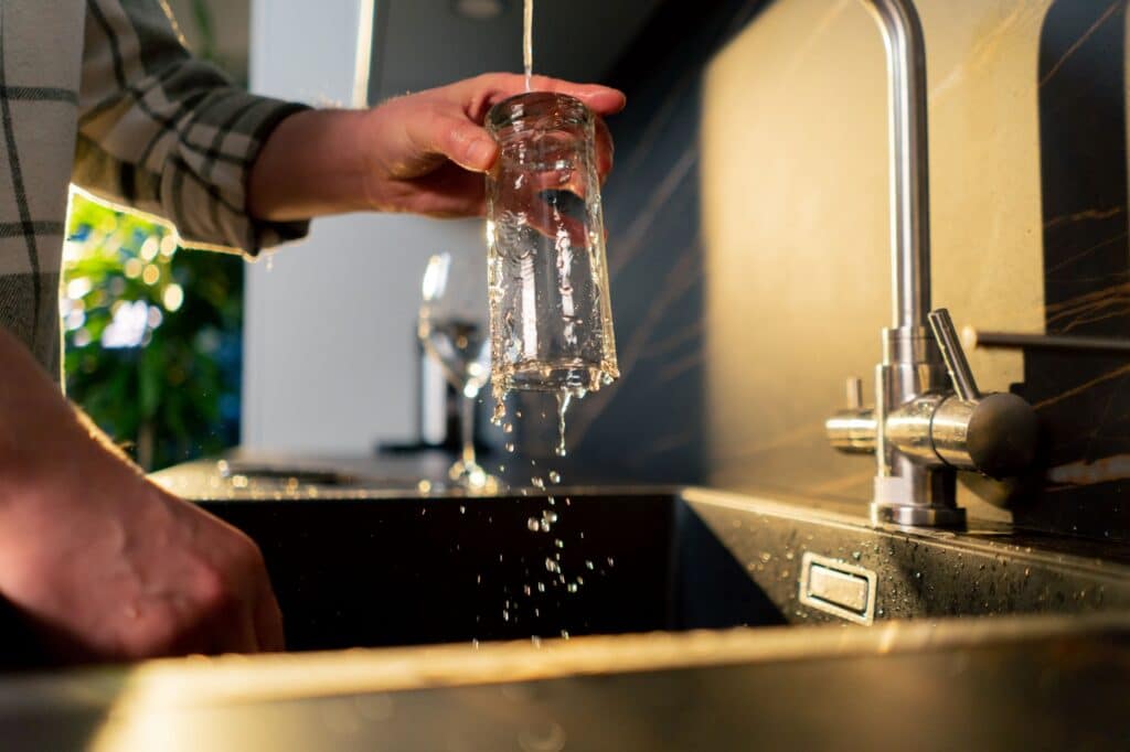A close-up shows a person's hand holding a clear glass upside down under a stream of water from a modern kitchen faucet, with water splashing into a dark sink.