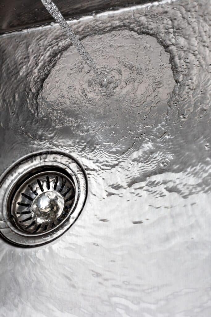 Clear water flows from a faucet into a stainless steel sink, creating circular ripples and bubbles as it drains through a silver metal strainer.