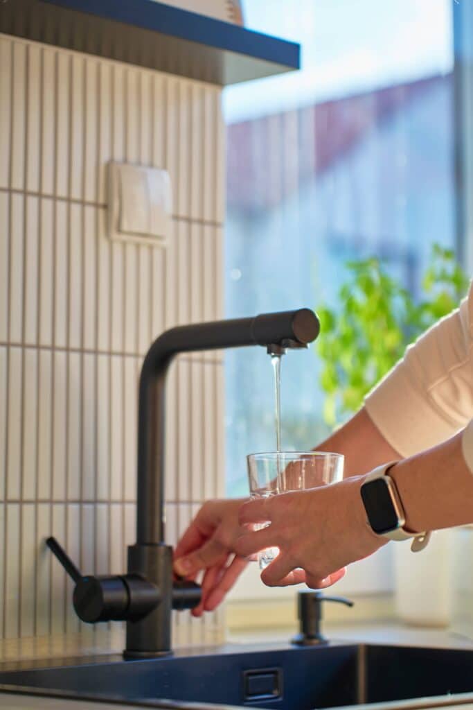 A person's hands fill a clear glass with water from a modern, matte black kitchen faucet against a background of white vertical tiles.