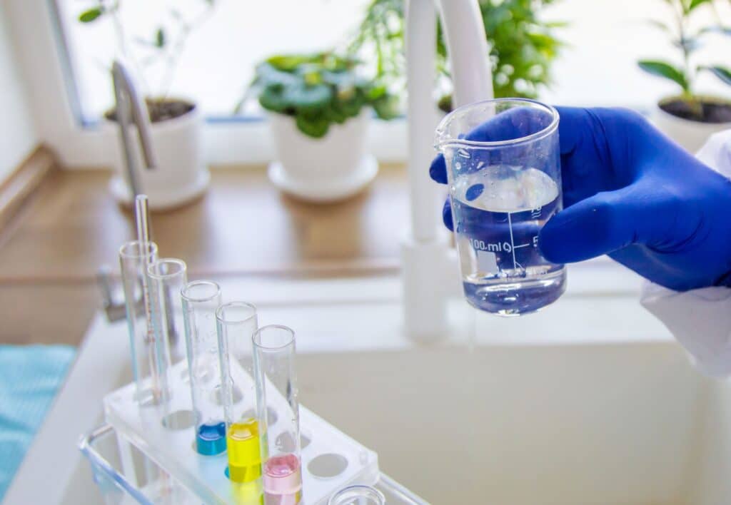 A person wearing a blue glove fills a glass beaker with water from a faucet next to a rack of colorful test tubes.