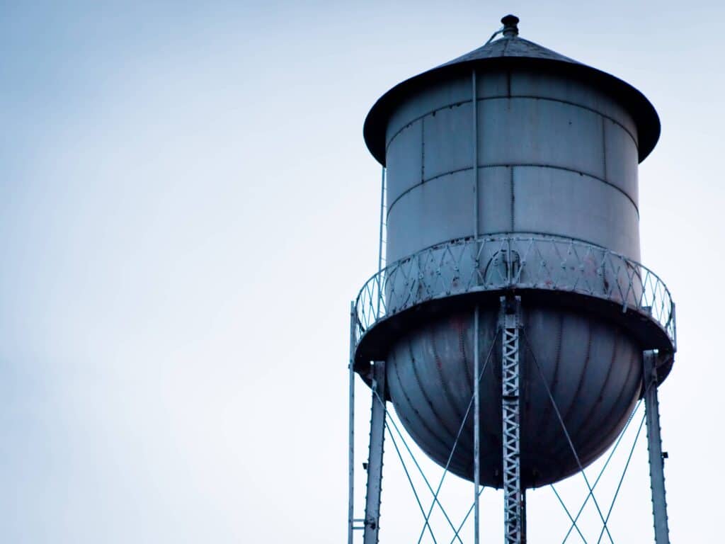 An old, weathered metal water tower stands tall against a pale blue sky, supported by a sturdy steel lattice frame.