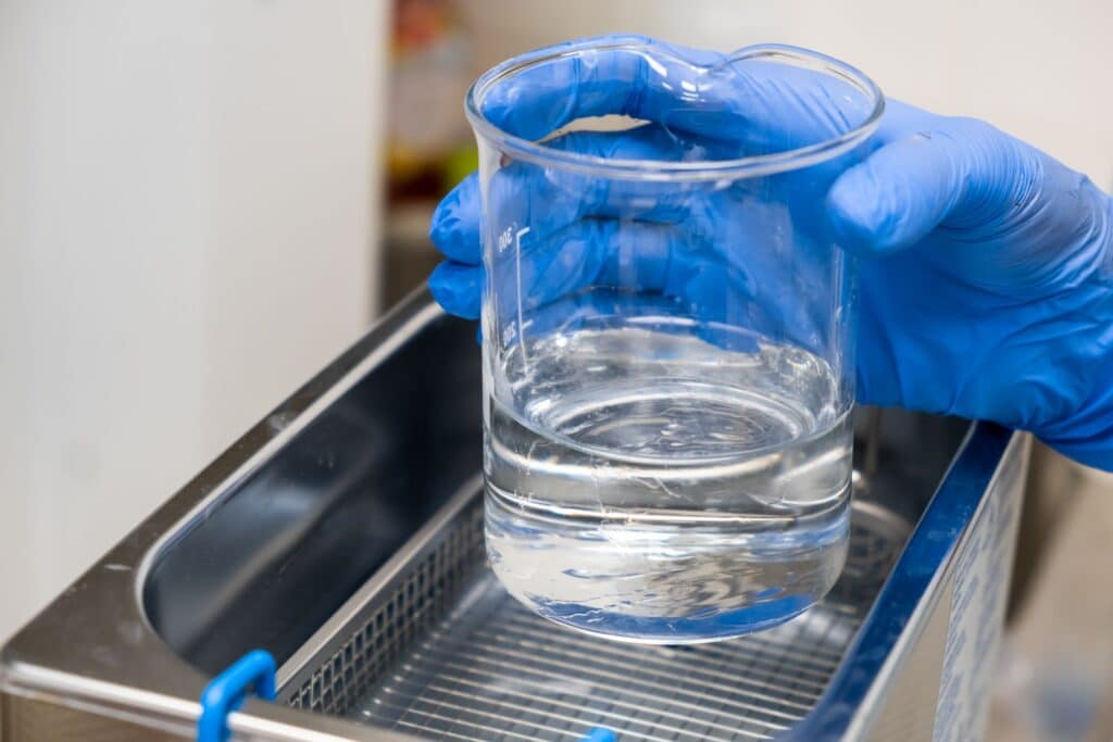 A person wearing blue nitrile gloves holds a glass beaker filled with clear liquid over a laboratory ultrasonic cleaner.