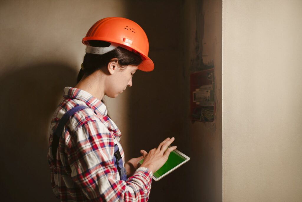 A home inspector wearing an orange hard hat and a plaid shirt uses a tablet with a green screen in a dimly lit indoor setting.