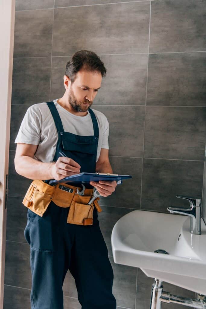 A focused plumber wearing blue overalls and a leather tool belt takes notes on a clipboard while standing in a modern bathroom next to a white pedestal sink.