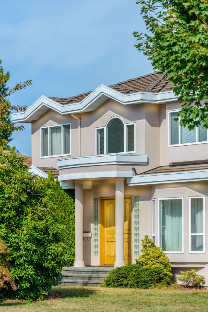 A modern two-story beige house featuring white columns flanking a golden front door, a distinctive zigzag roofline, and lush green landscaping under a clear blue sky.