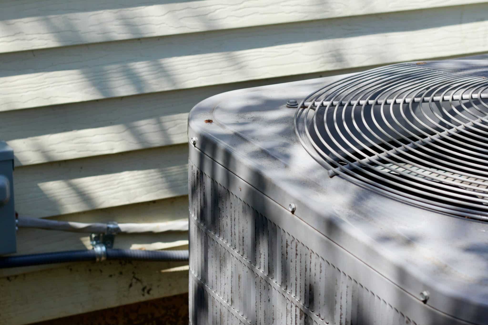 A close-up shot of an outdoor central air conditioning unit with a circular top grille, positioned next to the light-colored horizontal siding of a house with dappled shadows stretching across the surface. New Braunfels, TX